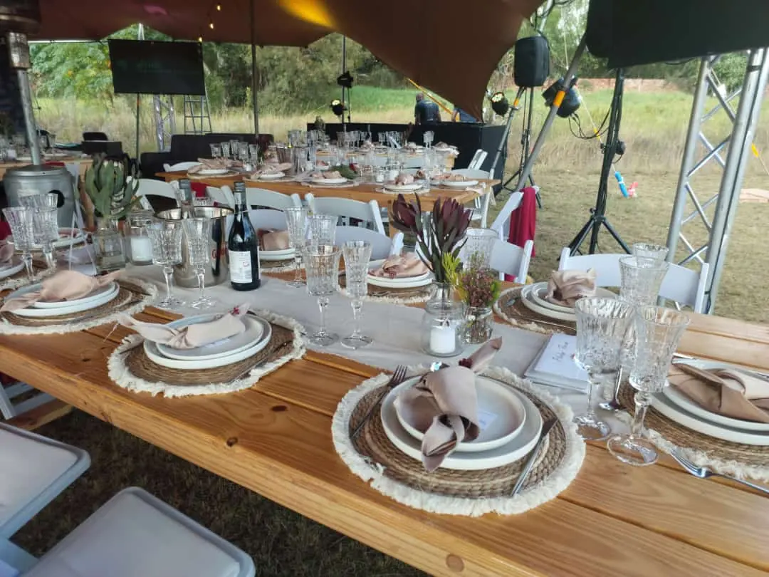 Full event equipment hire setup — long wooden trestle tables under a stretch tent with crystal glassware, charger plates, napkins, candles and stage lighting by Decorvent Hire Gauteng