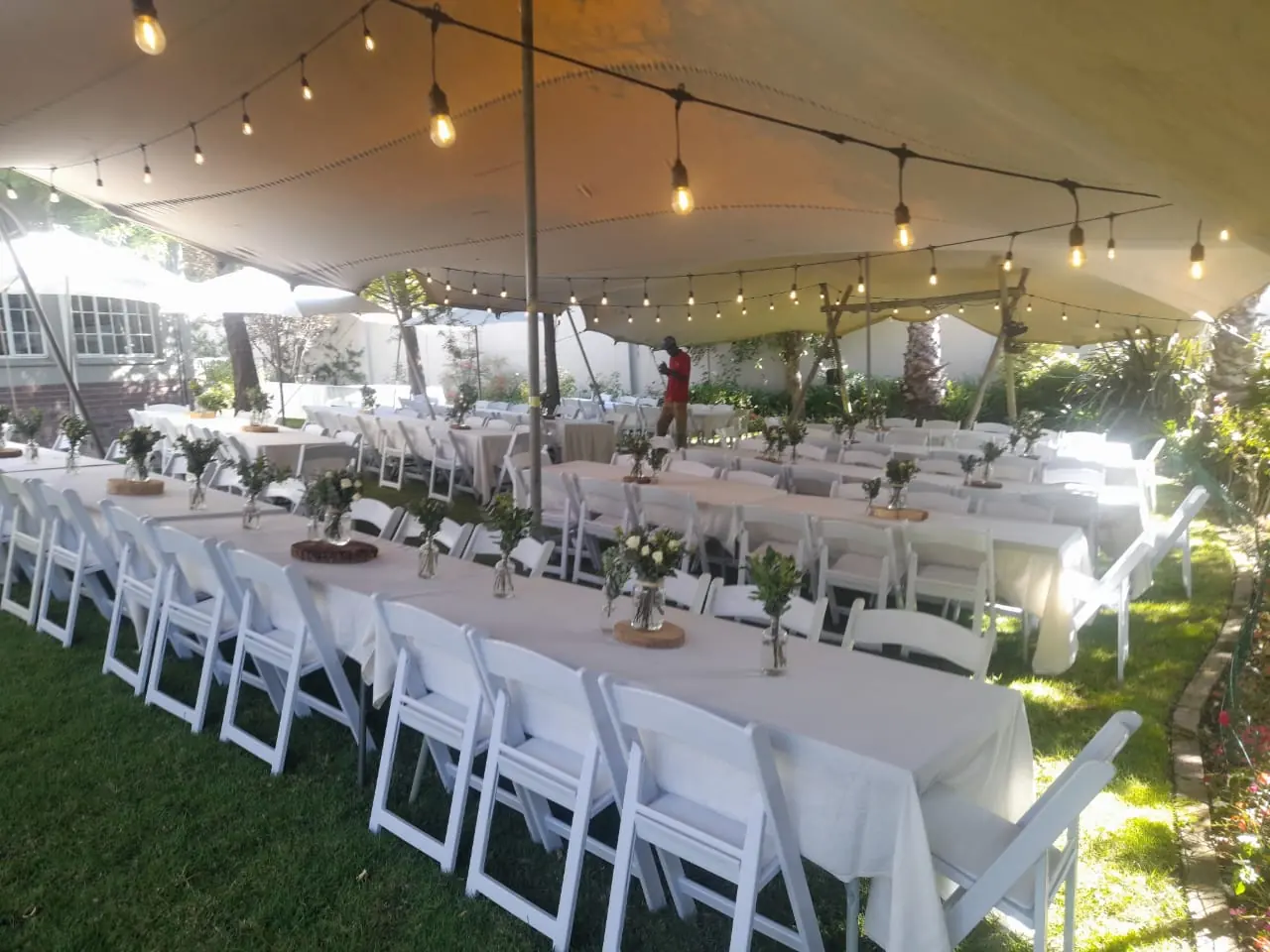 Long trestle tables with white folding Americana chairs and string lighting under a stretch tent — garden wedding table and chair hire by Decorvent Hire