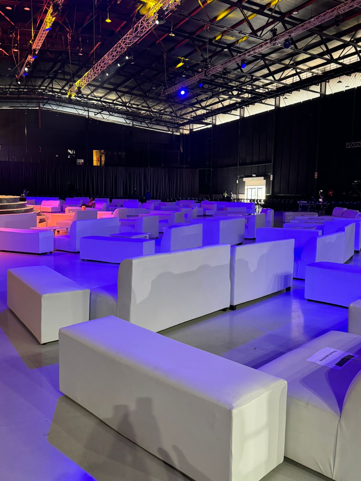 Rows of white lounge couches under blue and purple stage lighting at a large concert venue — VIP lounge furniture hire by Decorvent Hire
